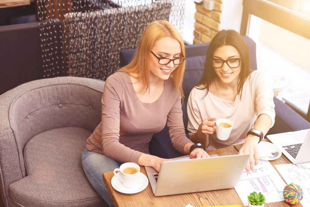Working together with joy. Pretty young woman is typing on laptop and laughing. Her friend is pointing finger at screen with inspiration. They are sitting and drinking coffee in cafe